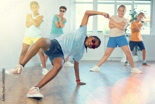 African boy in sun glasses performs choreographic exercises and teaches energetic mobile social dance Jazz-modern together with friends. Multi-racial group of children performs movements in studio