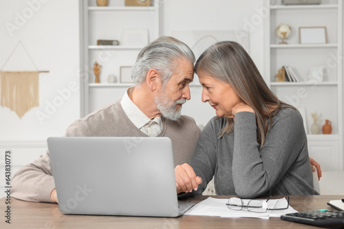 Elderly couple with papers and laptop discussing pension plan at wooden table in room