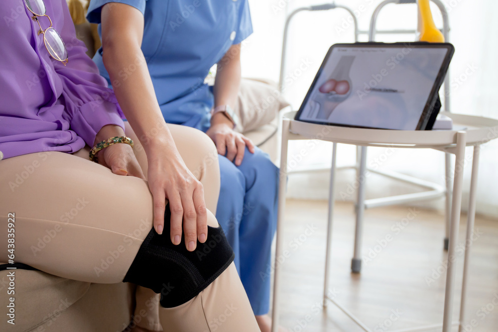 Obraz premium Caregiver or nurse helping check tendon and arthritis of knee or leg for diagnostic and rehabilitation while explaining senior woman with tablet living room at home, caretaker or physiotherapist.
