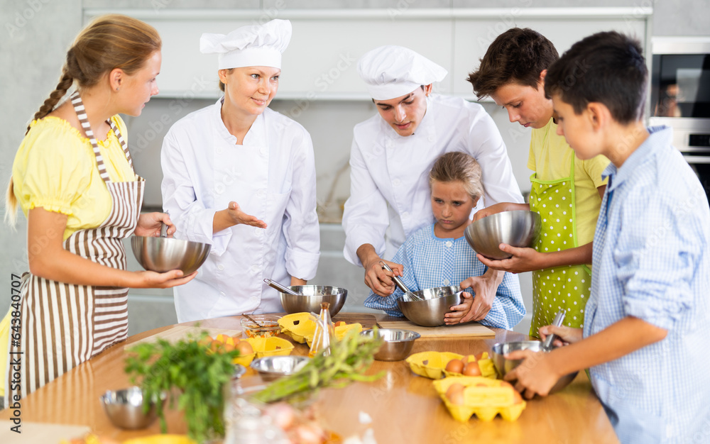 During lesson in cooking courses, female cook professional tells children about rules ticks and tricks for making fluffy pancakes, guy assistant helps hesitant child mix ingredients in bowl