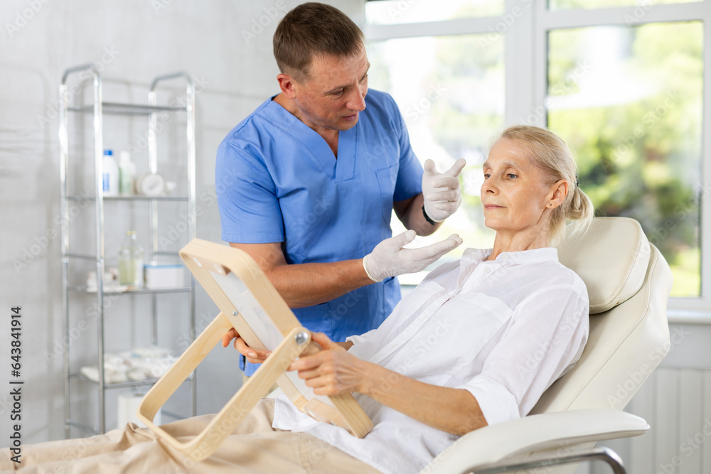 Fototapeta premium Beautician talking with patient before injection treatment, while client observing herself in mirror.