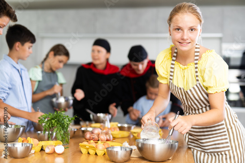 During cooking classes  girl carefully mixes ingredients in bowl with whisk. In background  blurry children stand near kitchen table and listen to chefs explanations