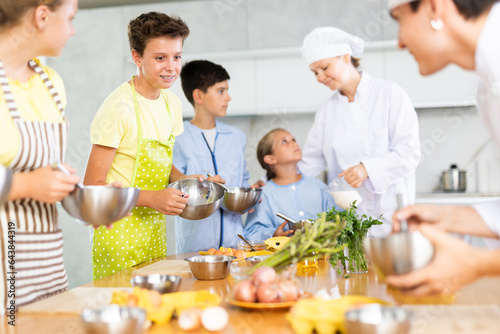 Young guy and adult woman cook in uniform teaches group of children how to cook dish