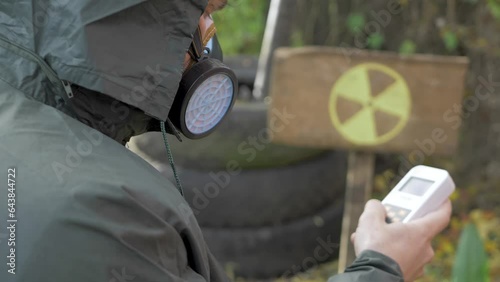 Use radiation geiger counter, checks level of radiation. Yellow danger sign in radioactive zone background. Nuclear weapons, atomic apocalypse concept. Male in protective suit and mask respirator  photo