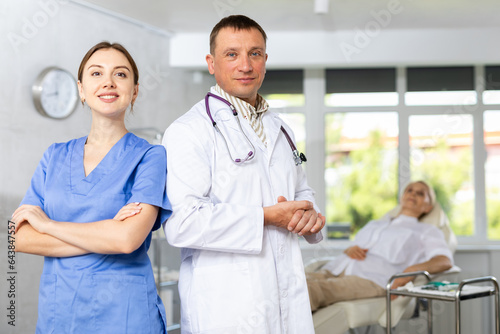 Positive woman and man medical specialists in uniform are standing in hospital. Doctor and nurse are waiting for procedure. Blurry patient in background