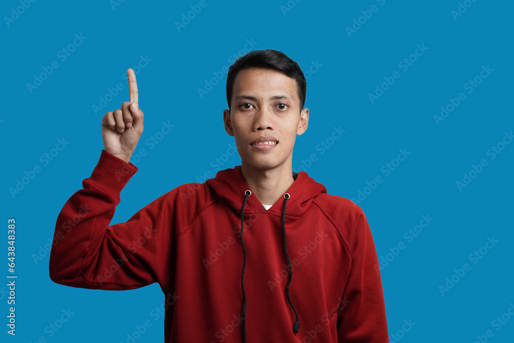 Excited young Asian man wearing red hoodie pointing up copy space, showing place for your ad isolated over blue background. Excited young Asian man wearing red hoodie pointing up copy space, showing place for your ad isolated over blue background.