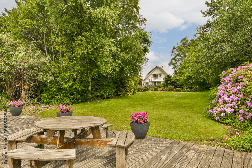 a picnic table and flowers on a wooden deck in front of a house with green grass, trees and blue sky a picnic table and flowers on a wooden deck in front of a house with green grass, trees and blue sky