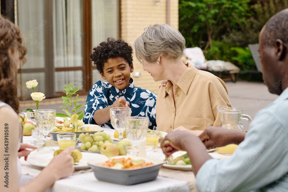 Cute smiling African American boy pointing at empty glass while talking to his grandmother by table served with homemade food and drinks