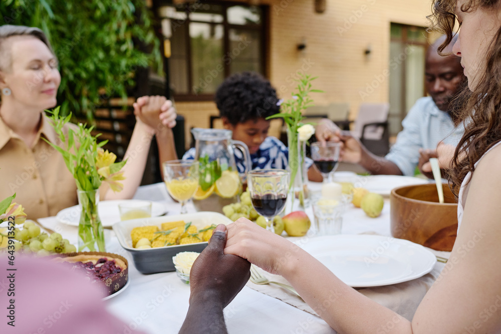 Focus on hands of young intercultural couple blessing food on served table before having outdoor dinner with their family members