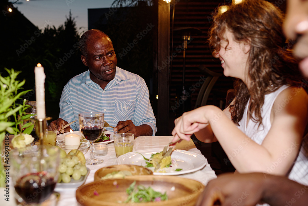 Young brunette woman sitting next to mature African American man by table served with homemade food, eating and listening to him by dinner Young brunette woman sitting next to mature African American man by table served with homemade food, eating and listening to him by dinner