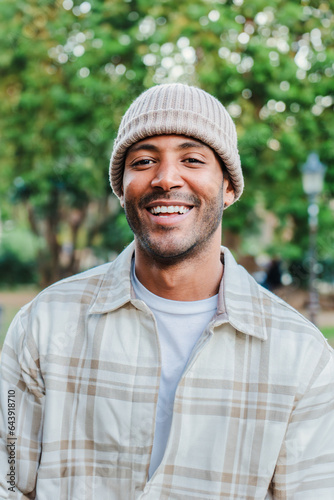 Vertical close up portrait of young hispanic man with a beanie hat smiling and looking at camera outdoors. Front view of latin happy guy standing at park with carefree attitude. Lifestyle concept