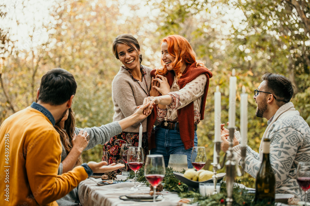 The joyous ambiance of a garden party, with a lesbian proposal with a diverse group of people relishing a shared meal amidst nature's beauty.