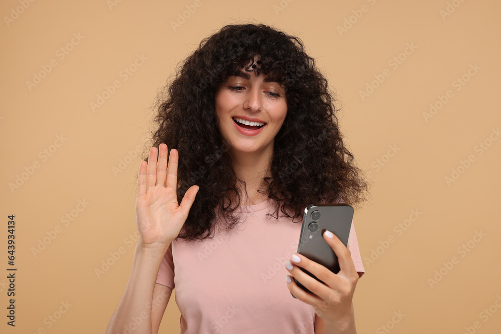 Happy young woman looking at smartphone and waving hello on beige background
