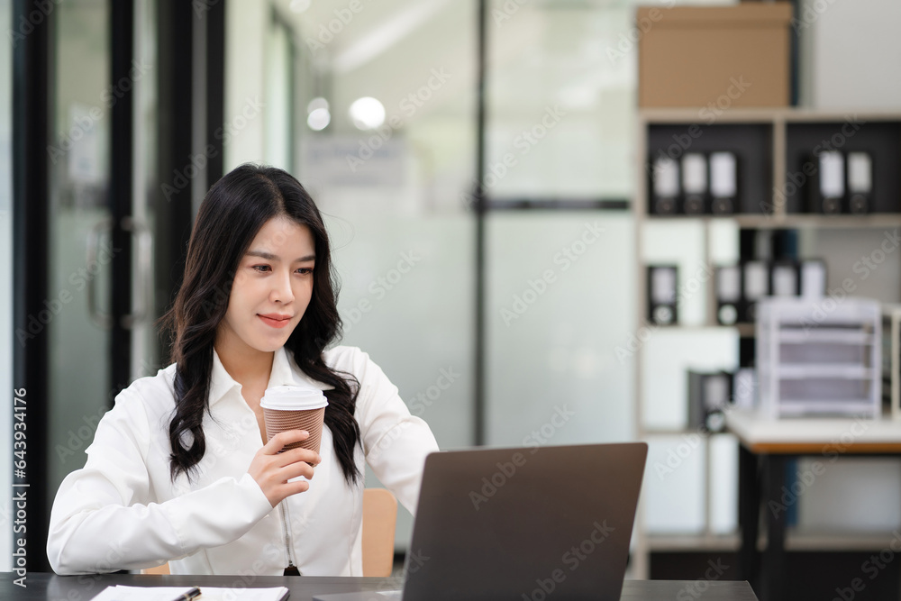 Asian businesswoman sits in a office working on laptop and enjoys a coffee. Asian businesswoman sits in a office working on laptop and enjoys a coffee.