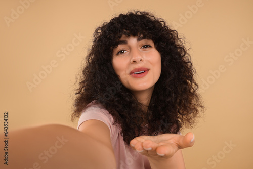 Happy young woman blowing kiss into camera and taking selfie on beige background