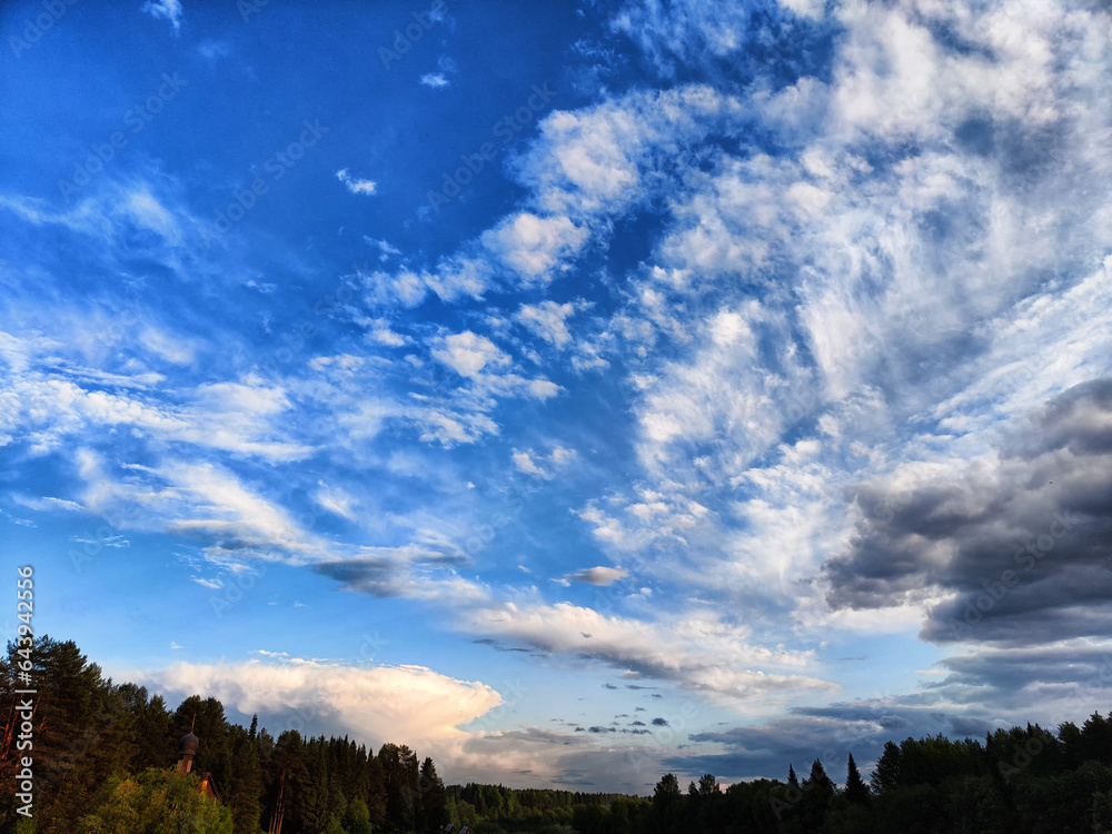 White fluffy clouds in the blue sky above the green trees in the forest. A beautiful natural landscape and scenery with clouds in the summer evening