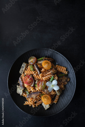 Fusilli pasta with mushrooms, asparagus and cherry tomatoes on a modern design plate with dark background