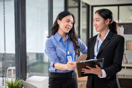 Two happy Asian businesswomen are having a discussion while standing in the office together.