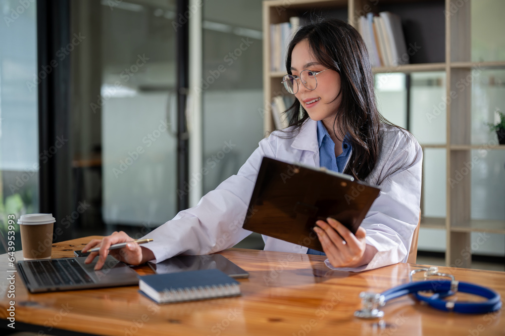 A professional Asian female doctor is working on her laptop at a table in her office at a hospital. A professional Asian female doctor is working on her laptop at a table in her office at a hospital.