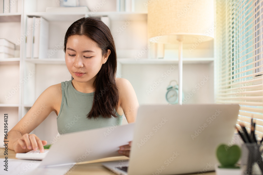 Asian woman working at home, Press the calculator to calculate the income - expenses of the house and use the laptop to record the information and check the accuracy, Home lifestyle. Asian woman working at home, Press the calculator to calculate the income - expenses of the house and use the laptop to record the information and check the accuracy, Home lifestyle.