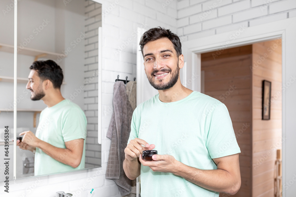 Handsome man looking at mirror and applying moisturizing cream on cheeks in bathroom. Groomed young guy doing skincare morning routine after taking a shower