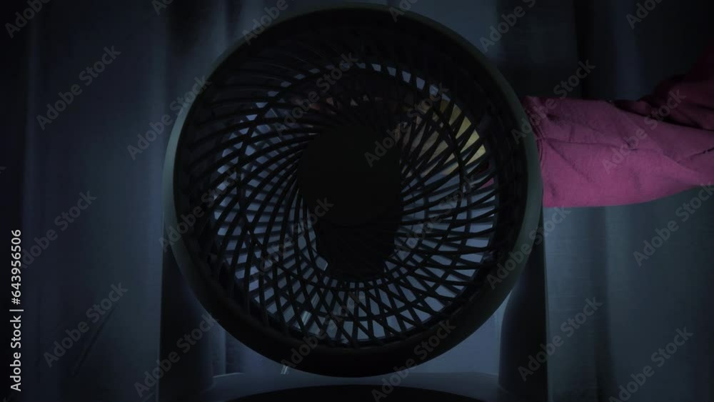 Closeup of a man’s hand turning off a small portable plastic cooling fan, with a blade propeller, in a darkened room with closed curtains.