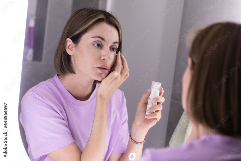 Attractive woman looking at mirror and applying moisturizing cream on cheeks in bathroom. Groomed young girl doing skincare morning routine after taking a shower