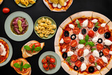 An exquisite spread of Italian cuisine featuring pizza, pasta, ravioli, carpaccio, caprese salad, and tomato bruschetta, elegantly arranged on plates. Top view on a dramatic black background
