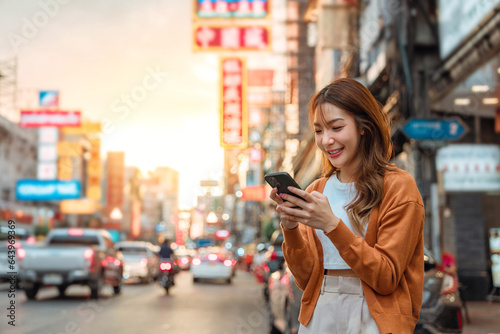 Happy young Asian tourist woman using smartphone on street with lights bokeh background, Female traveller in Yaowarat Chinatown street food market during sunset in Bangkok, Thailand. © oatawa