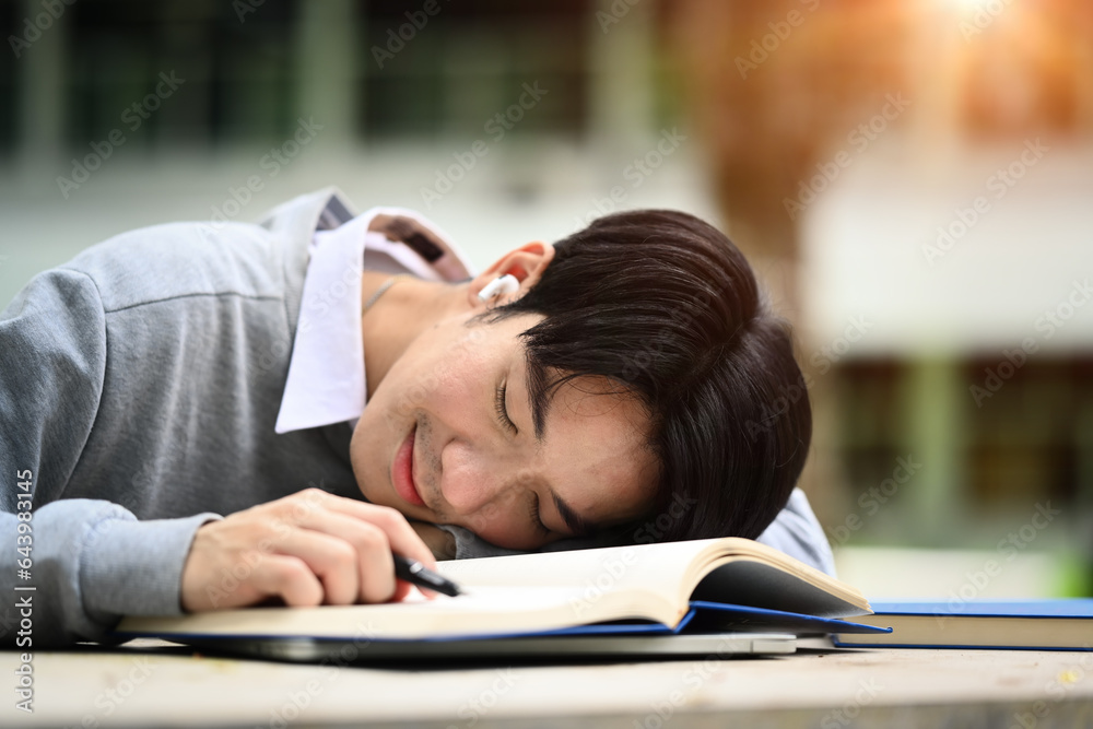 Asian male university student fallen asleep on his textbook, being exhausted during preparing for exams Asian male university student fallen asleep on his textbook, being exhausted during preparing for exams