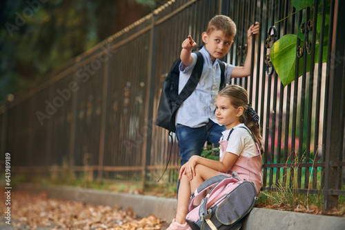By the fence, relaxing. Young school children of boy and girl are together outdoors