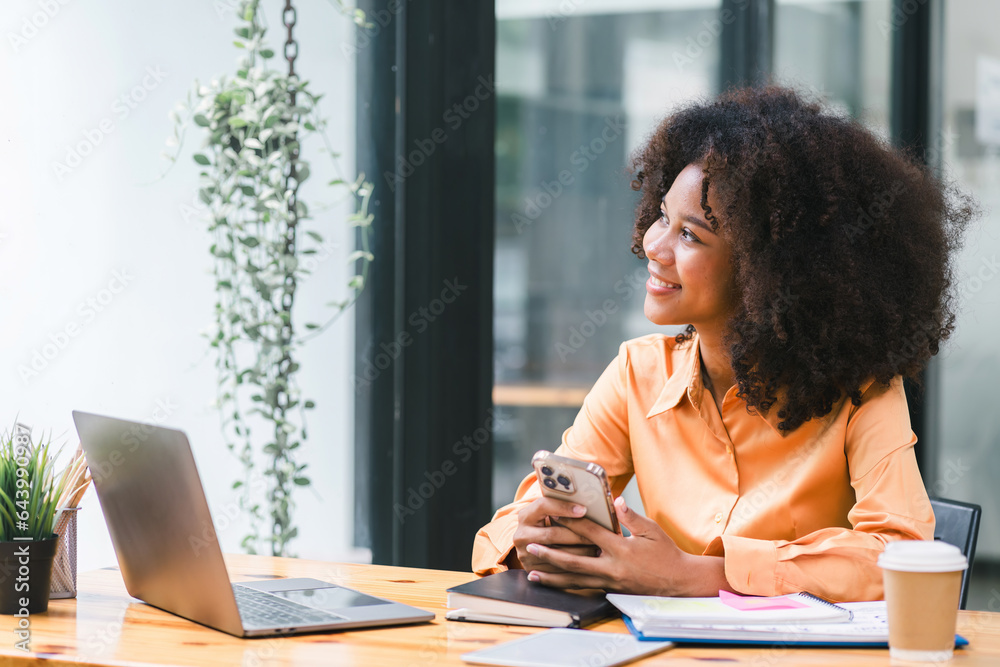 Cheerful young African American pretty woman, entrepreneur in casual wear, smiling, using smartphone and working in a modern office.