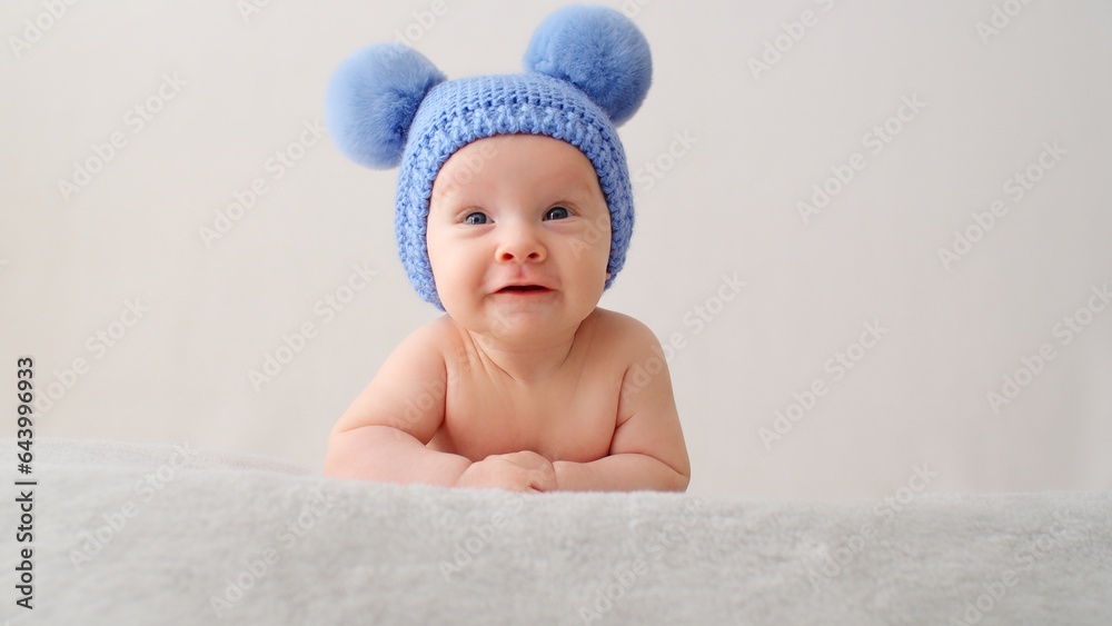 A smiling infant baby in a blue hat with pompoms looks into the camera. A happy newborn is lying on a blanket. Beautiful, happy little child with blue eyes.  Cheerful White kid.