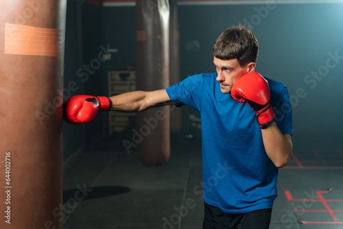 Tall white man in a blue tee shirt engages in a focused boxing punching exercise with a heavy bag, showcasing his dedication to boxing and fitness in a boxing gym © asean studio