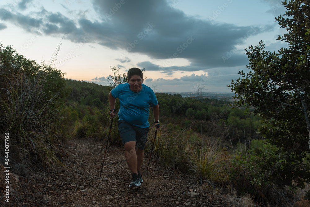 Overweight man trains in the mountains supported by sports poles.