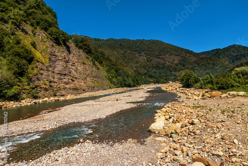 Landscape with mountains, forest and river in the foreground. The mountain river flows through a rocky bed. A mountain river flows near the forest on a sunny day. A river in the mountains.