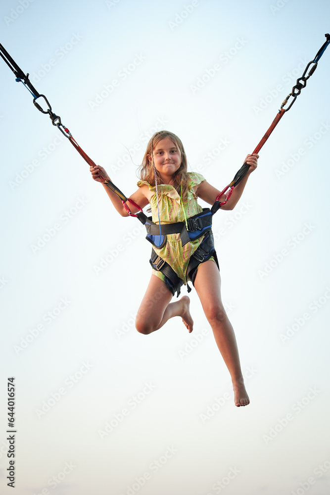 Bungee jumping at trampoline. Little girl bouncing on bungee jumping in amusement park on summer vacations. Super girl