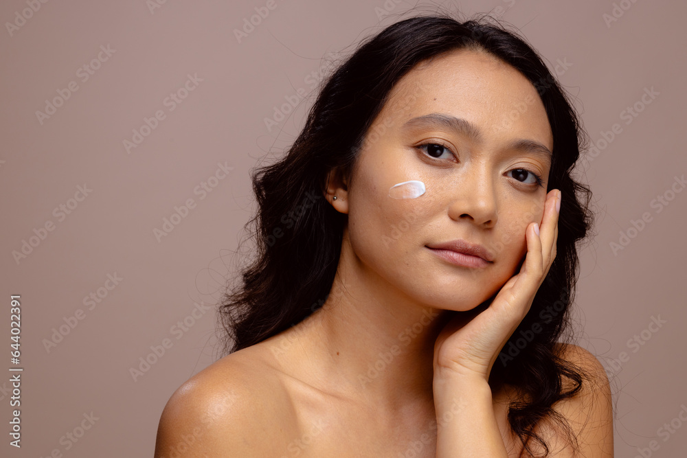 Asian woman with dark hair with skin cream on her cheek touching face with hand, copy space Asian woman with dark hair with skin cream on her cheek touching face with hand, copy space