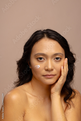 Asian woman with dark hair with skin cream on her cheek touching face with hand, copy space