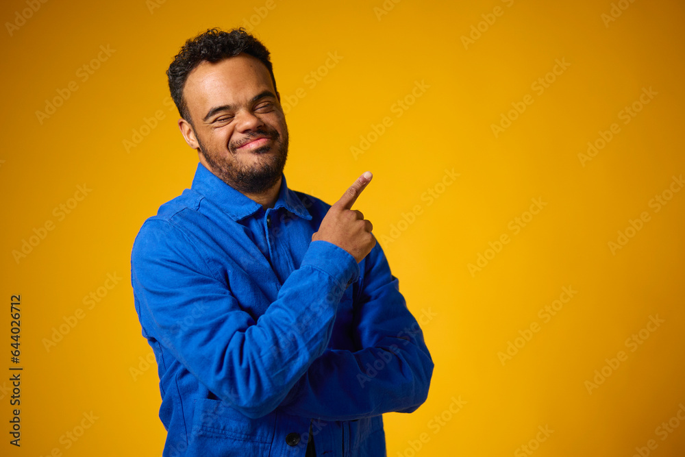 Studio Portrait Of Smiling Man With Down Syndrome Standing Against Yellow Background Pointing