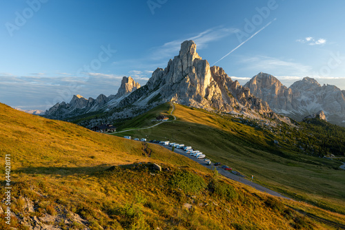Sunrise at Passo Giau with road and parked cars and campers with sun shining at meadow and peaks in  the background during summer morning in august and grass in foreground