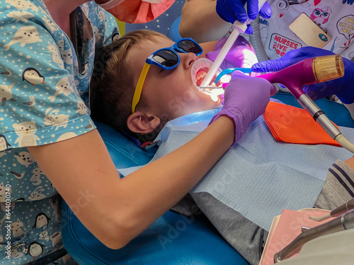 female dentist and nurse treating little boy patient with lip and cheek retractor on his mouth sitting in chair at dental office