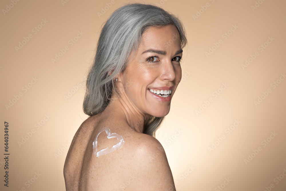 Obraz premium Attractive senior woman with heart made of body cream on back standing half-turned and smiling, free space