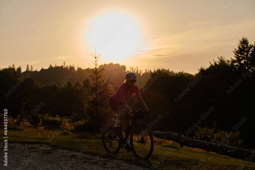 Obraz premium A fit female cyclist, wearing a cycling kit and helmet, is riding a gravel bike on a gravel road at sunset. Sports motivation image.
