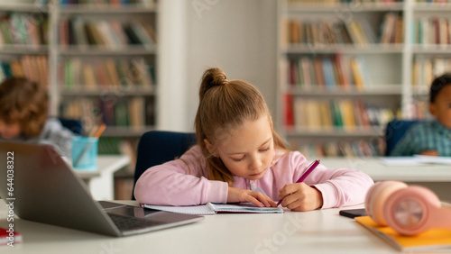 Smart schoolgirl taking notes while e-learning on laptop computer, sitting at desk with classmates on background
