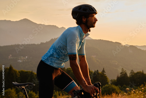 Man cyclist wearing blue cycling jersey. He is riding a gravel bike on a gravel road at sunset with a view of the mountains.Empty mountain road.Cycling gravel adventure in Romania.Bucegi Natural Park photo