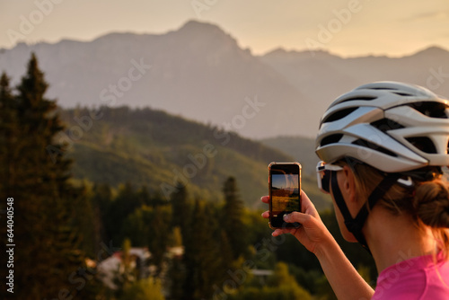 Female cyclist wearing cycling kit and helmet ,pauses to take a photo breathtaking mountain scenery on her mobile phone during training. Bucegi Mountains, Romania photo
