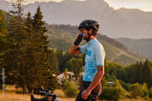 Man cyclist in mountain terrain quenches his thirst during intense training. Male cyclist in cycling kit and a helmet is going to drink water from a sports bottle. Bucegi Mountains, Romania photo