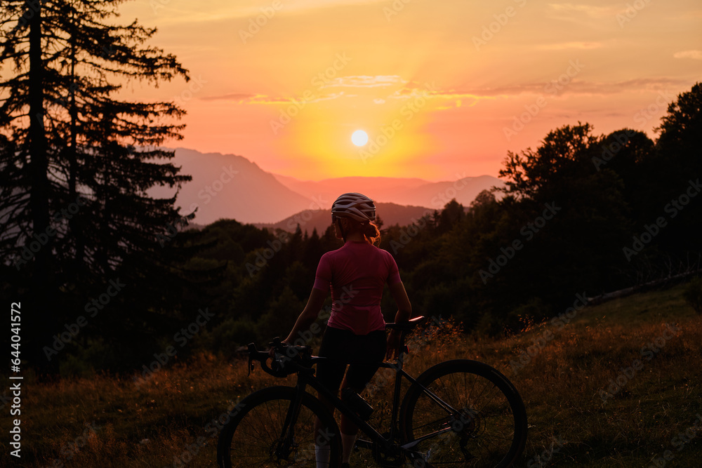 Fototapeta premium A woman cyclist on a bicycle is enjoying the sunset in the mountains from a back view. Beautiful evening landscape with a bicycle. 