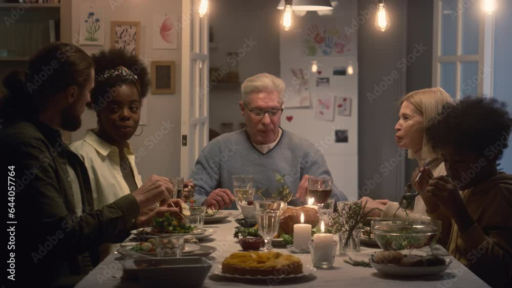 Multi-ethnic family of five multi-generational members talking to each other while enjoying food sitting at table Multi-ethnic family of five multi-generational members talking to each other while enjoying food sitting at table